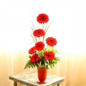 Florero rojo en forma de copa con 7 gerberas rojas.