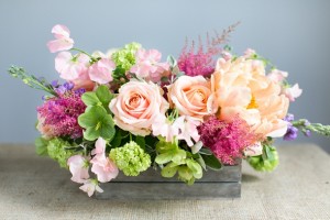 Arreglo de flores primaverales en tonos rosados con follaje verde en bandeja.